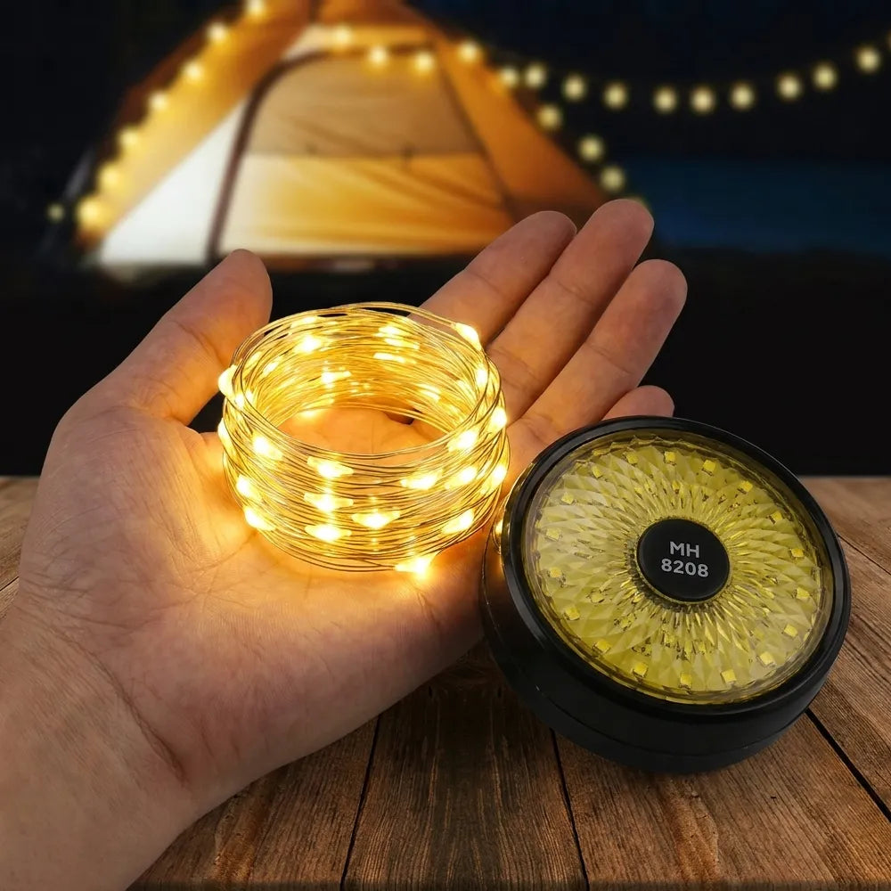 A hand holds a coiled bundle of warm white LED fairy string lights next to a round black container lid with a yellow glittery insert and a label reading MH 8208, set against a blurred nighttime camping scene with a tent and string lights.