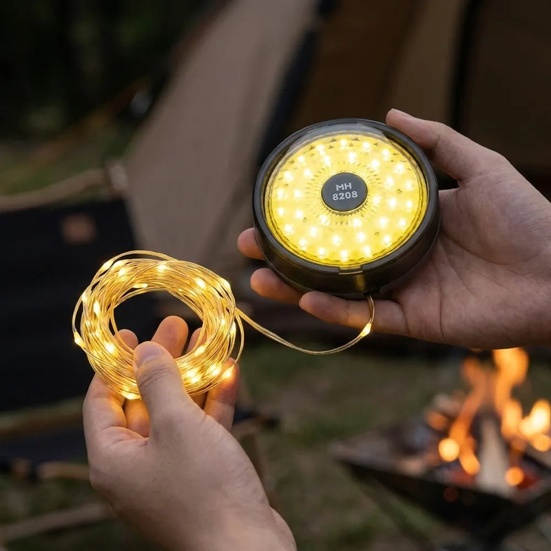 A person holds a round solar LED light and a coiled string of warm white fairy lights outdoors near a campfire, with a tent blurred in the background.