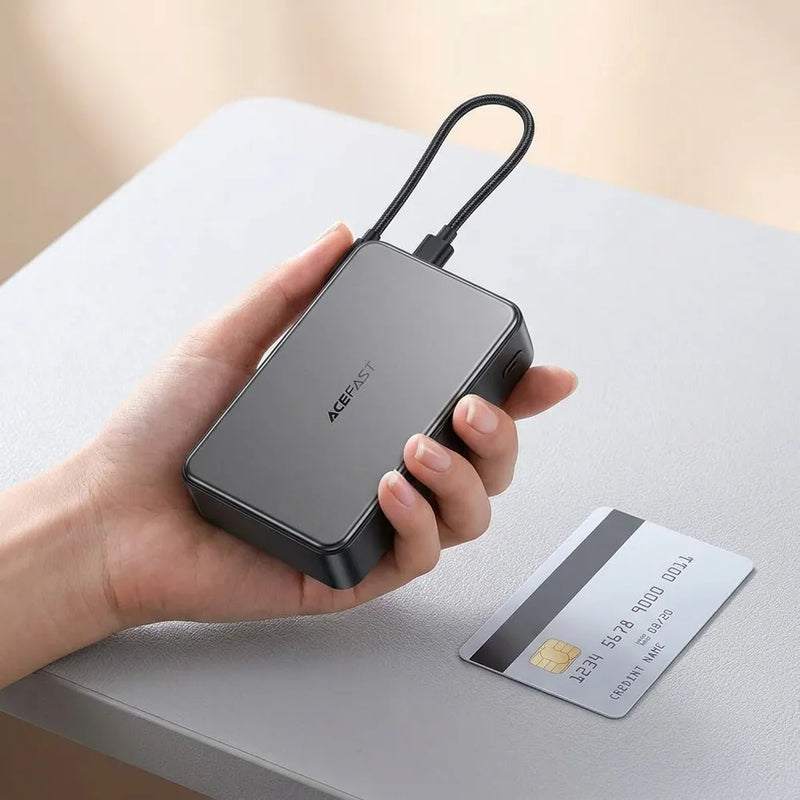 A hand holds a compact black rectangular electronic device with a looped cable, branded ACEFAST, next to a credit card on a light gray surface.