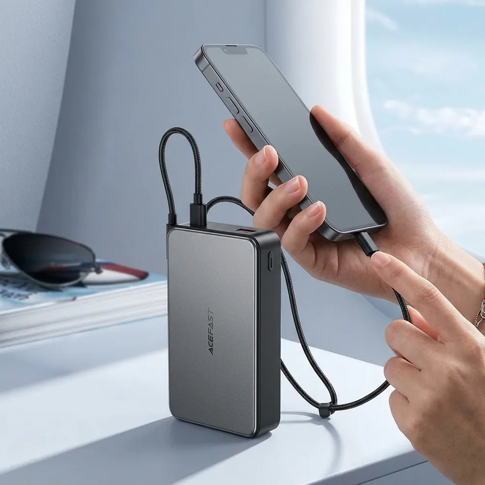 A portable gray power bank with a black cable is shown being used to charge a smartphone, with a person's hands holding the phone and pointing at the cable. The background is a bright, modern setting with a window view.