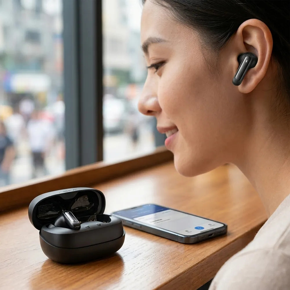 A woman wearing a dark gray true wireless earbud is shown in profile next to an open black charging case and a smartphone on a wooden table.