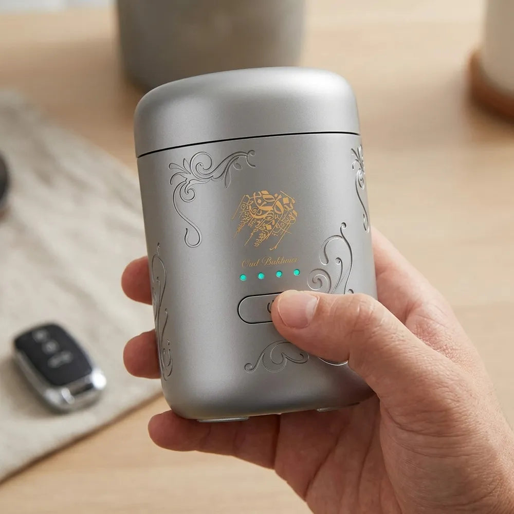 A hand holding a silver cylindrical electric kettle with gold decorative engravings and a gold lion emblem. The background shows a light wood table with a blurred key fob.