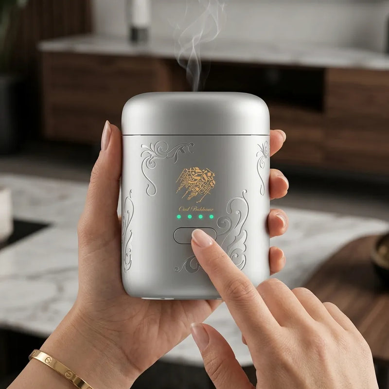 A silver handheld diffuser or humidifier with gold decorative engravings and a gold bee logo, held in two hands. Steam is visibly rising from the top, and a finger is pressing the front button.