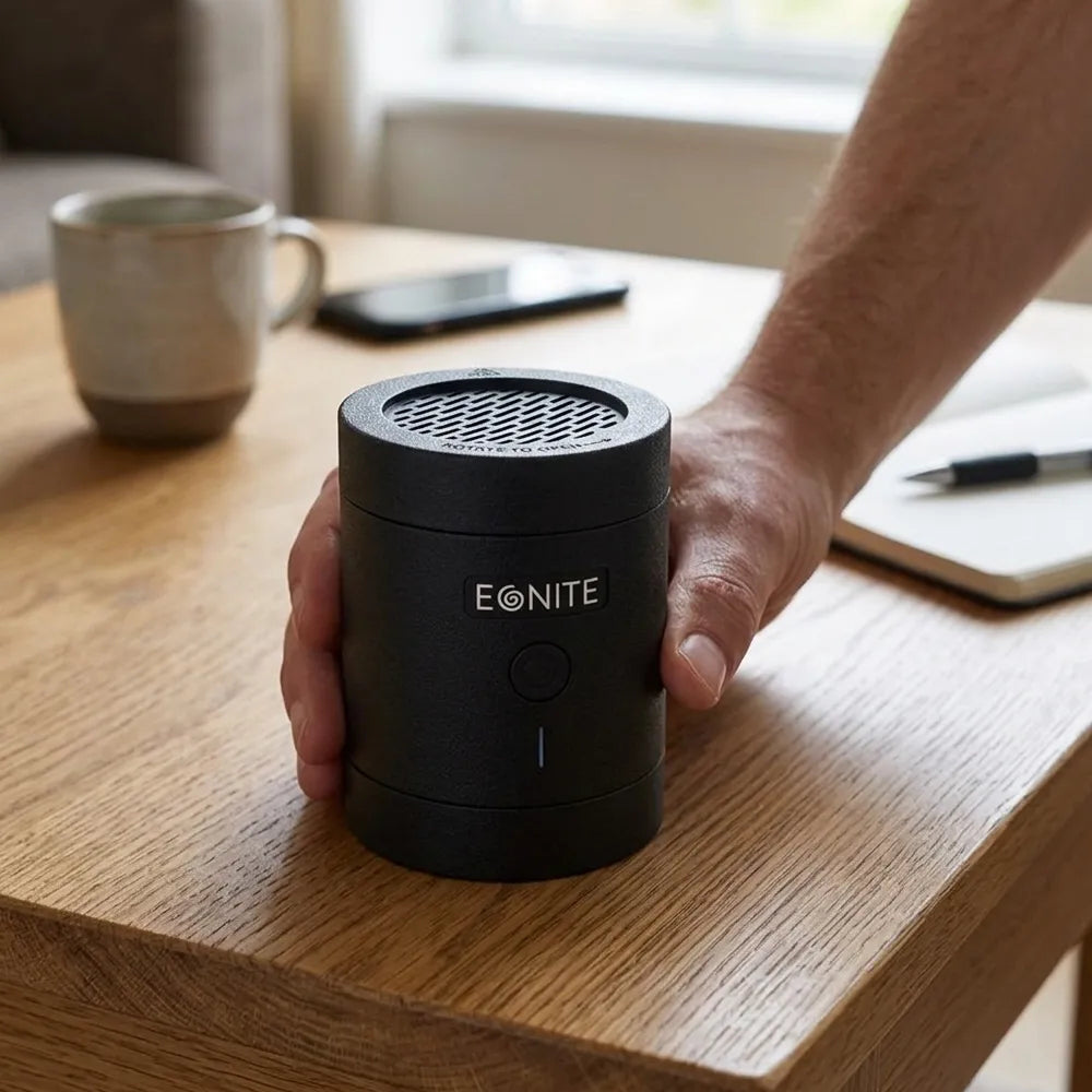 A person's hand holding a black cylindrical EONITE portable speaker on a wooden table, with a mug, phone, and notebook in the background.