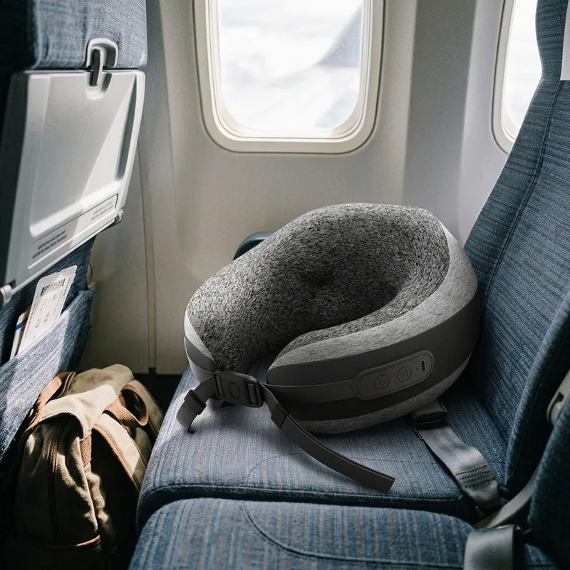 A gray and black neck pillow resting on a blue airplane seat next to a window, with an airplane tray table and a brown backpack visible in the cabin.