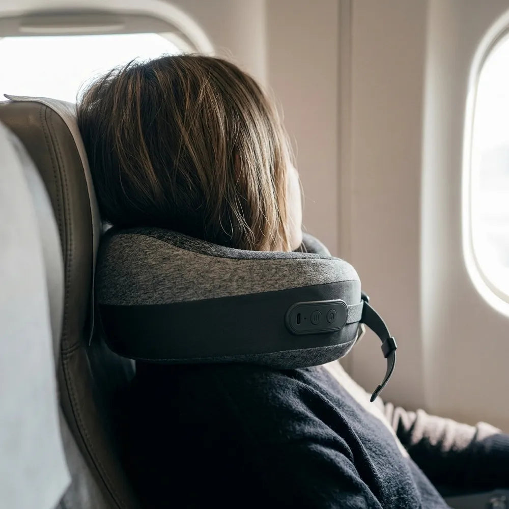 A person seated in an airplane cabin wearing a gray and black neck pillow around their head, with the window visible on the right.