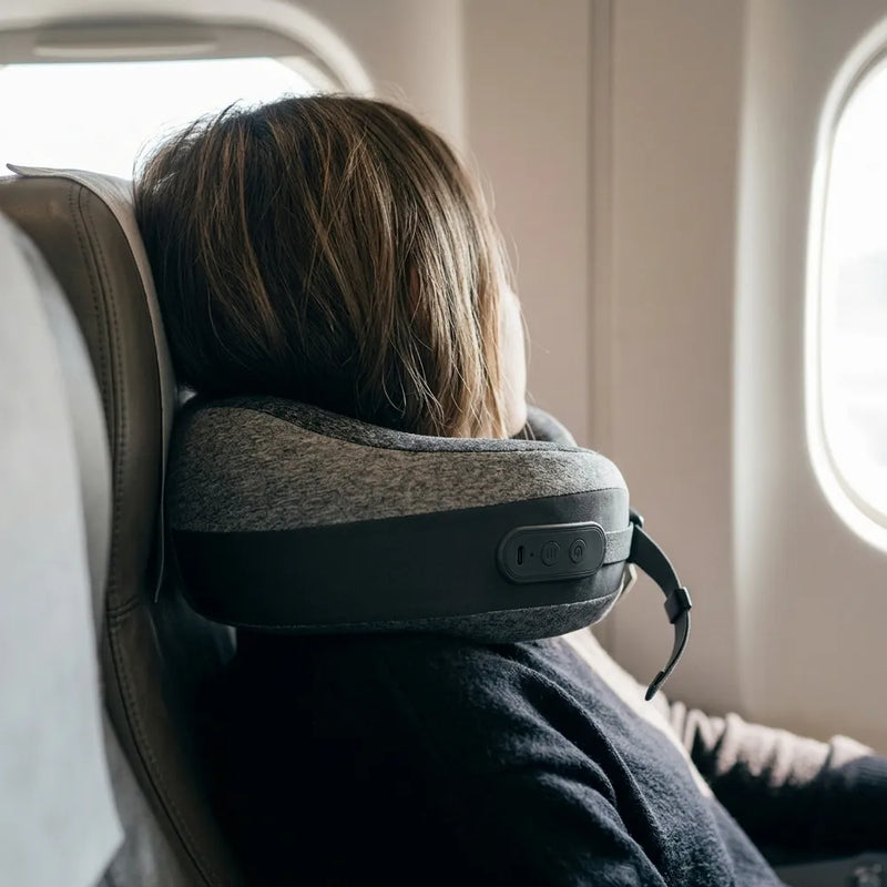 A person seated in an airplane cabin wearing a gray and black neck pillow around their head, with the window visible on the right.