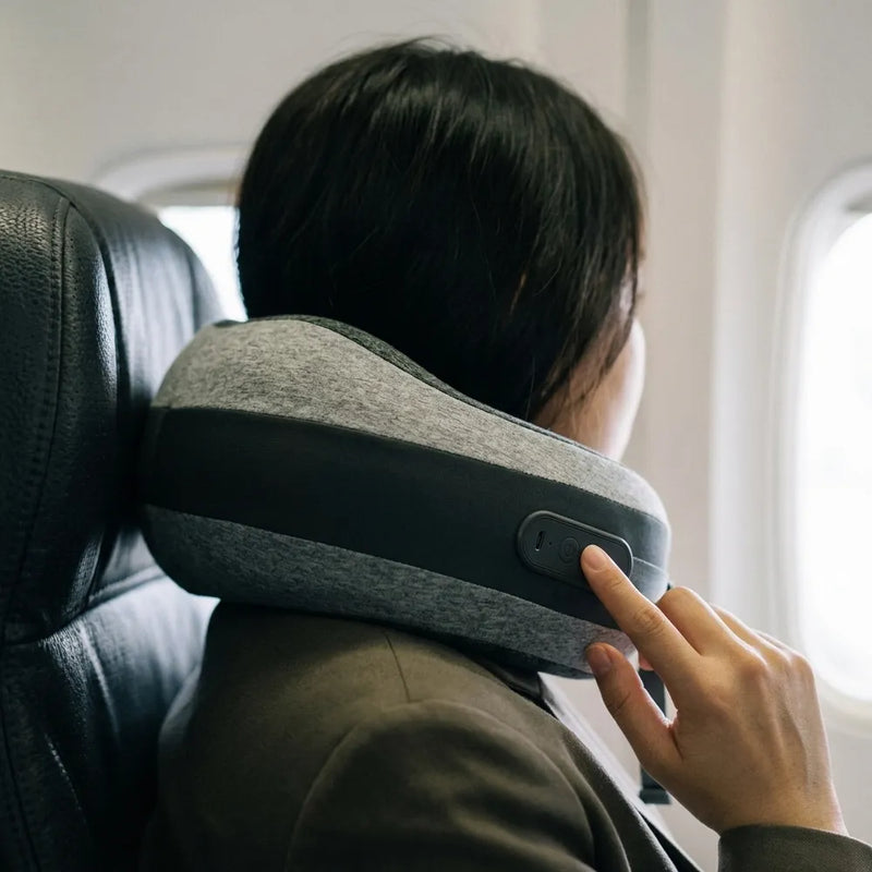 A person seated in an airplane cabin wearing a gray and black neck pillow around their neck, with their hand touching a control on the side of the pillow.