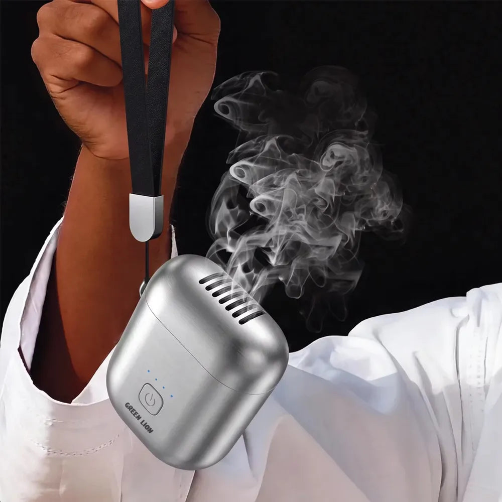 A silver portable air freshener diffuser with a black wrist strap, shown emitting white smoke. It is held by a person wearing a white shirt against a solid black background.