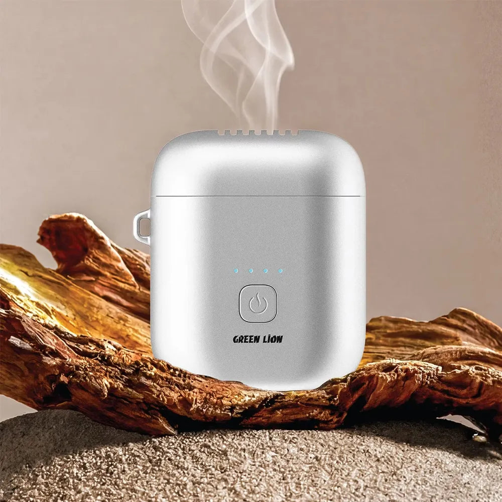 A silver electric humidifier with visible steam rising from the top, placed on a piece of driftwood over sand. The device features a power button and the brand name 'GREEN LION' printed on the front.