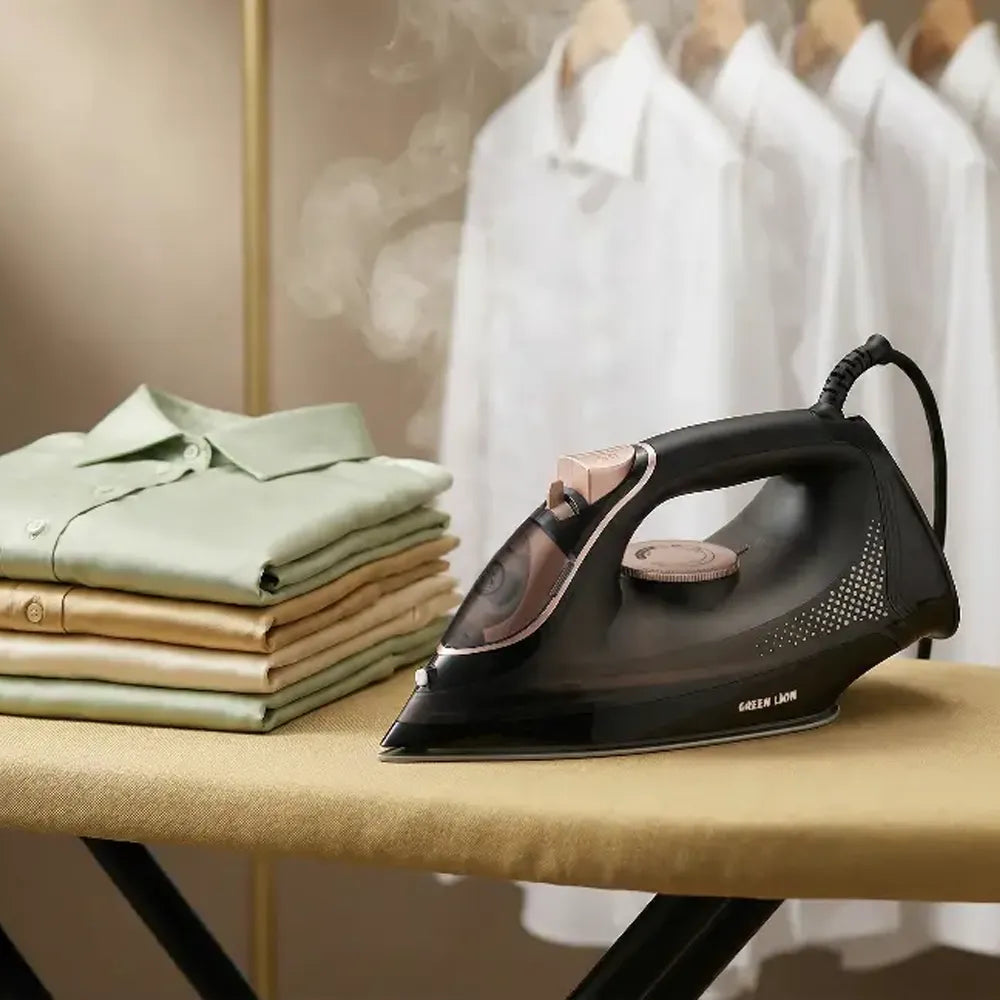 A black steam iron with rose gold accents sits on a beige ironing board, with folded green and beige shirts on one side and white shirts hanging in the background.