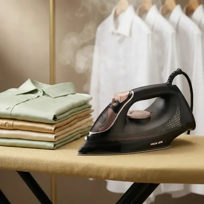 A black steam iron with rose gold accents sits on a beige ironing board, with folded green and beige shirts on one side and white shirts hanging in the background.