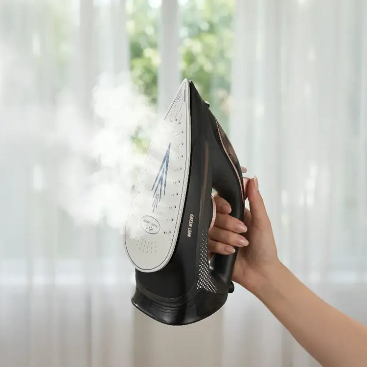 A hand holding a black and silver steam iron that is emitting a visible plume of steam, set against a soft-focus background of white curtains and green foliage.