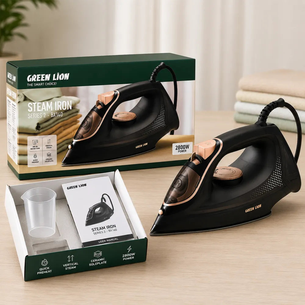 A Green Lion steam iron shown alongside its retail box and included accessories on a light wood tabletop. The iron is black with rose-gold metallic accents, and the packaging is green, white, and tan.