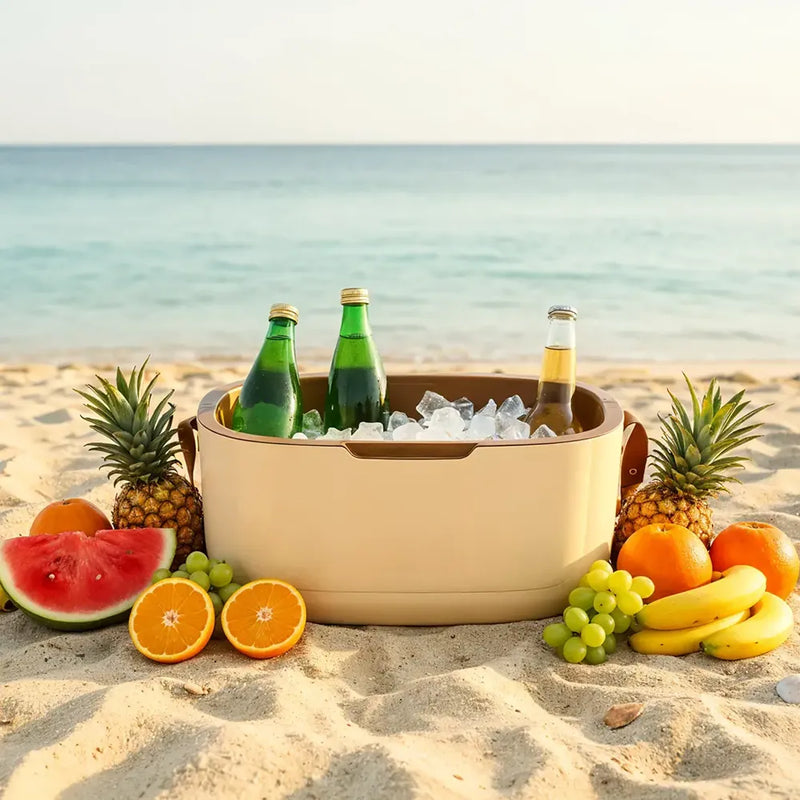 A beige outdoor ice bucket filled with ice and three beverage bottles, set on a sandy beach with water in the background. Fresh fruit including watermelon, oranges, grapes, bananas, and pineapples is arranged around the bucket.