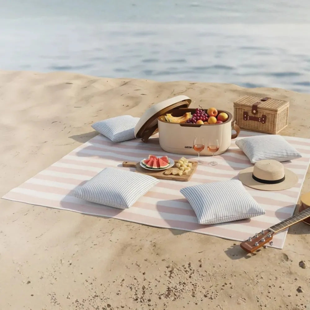 A beach picnic setup on a striped blanket with four blue-and-white striped pillows, a cream electric kettle filled with fruit, a woven storage box, a straw hat, and a guitar on a sandy shore by the water.