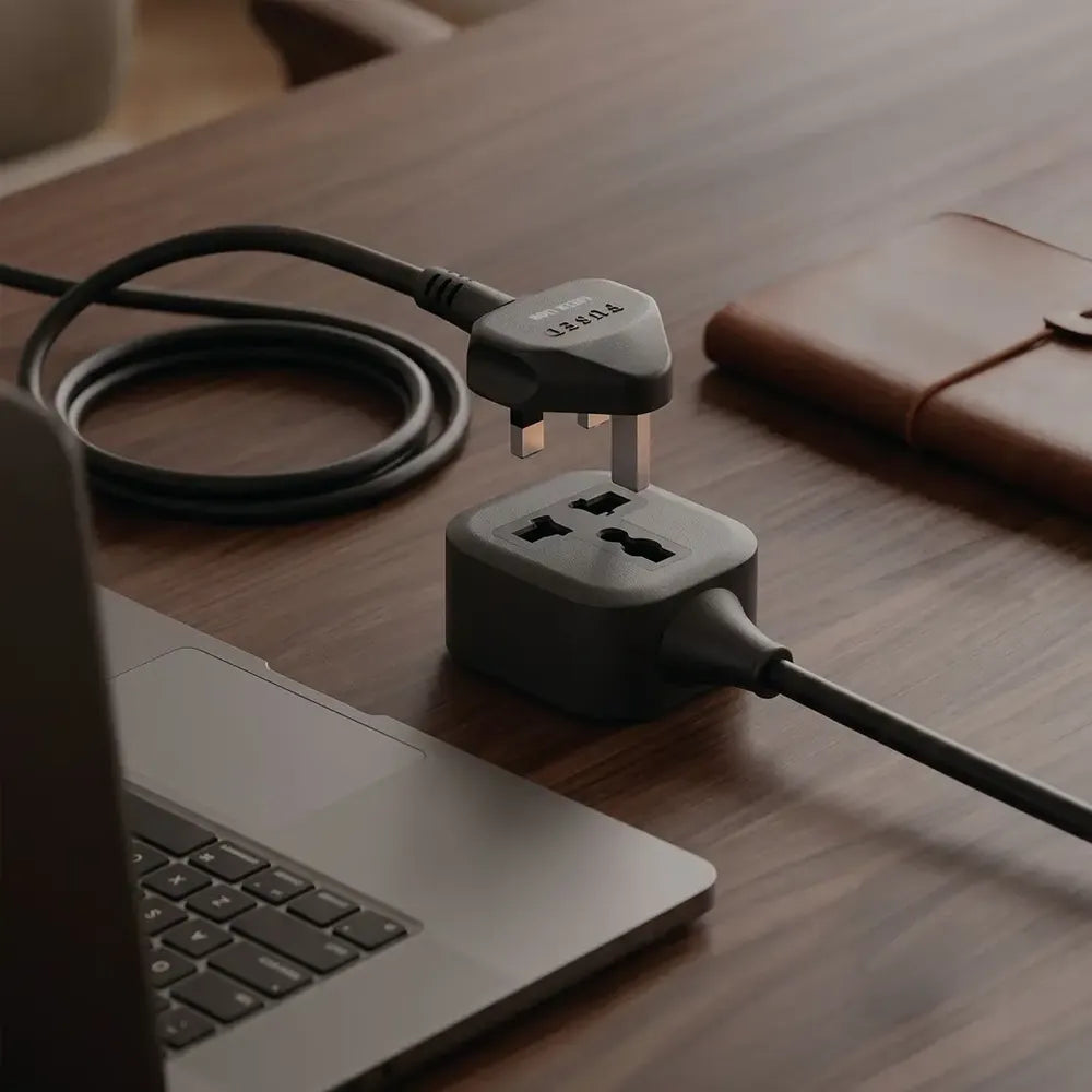 A dark gray multi-socket power adapter with a UK three-pin plug inserted, shown on a wooden desk next to a laptop and a brown leather notebook.