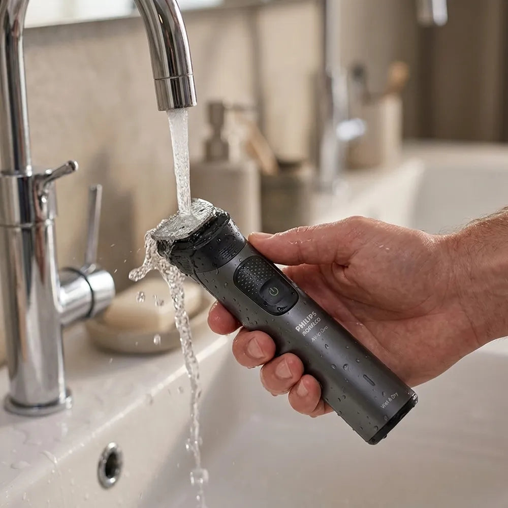 A hand holding a dark gray electric shaver under running water from a chrome faucet in a bathroom setting.