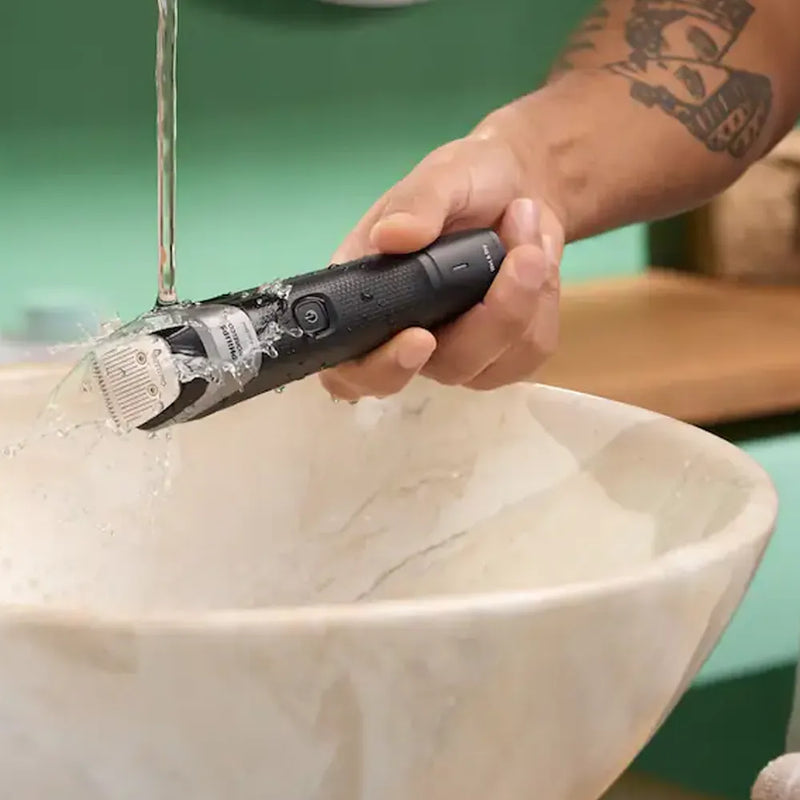 A close-up of a person's tattooed arm holding a black electric beard trimmer under a running water stream above a marble sink.