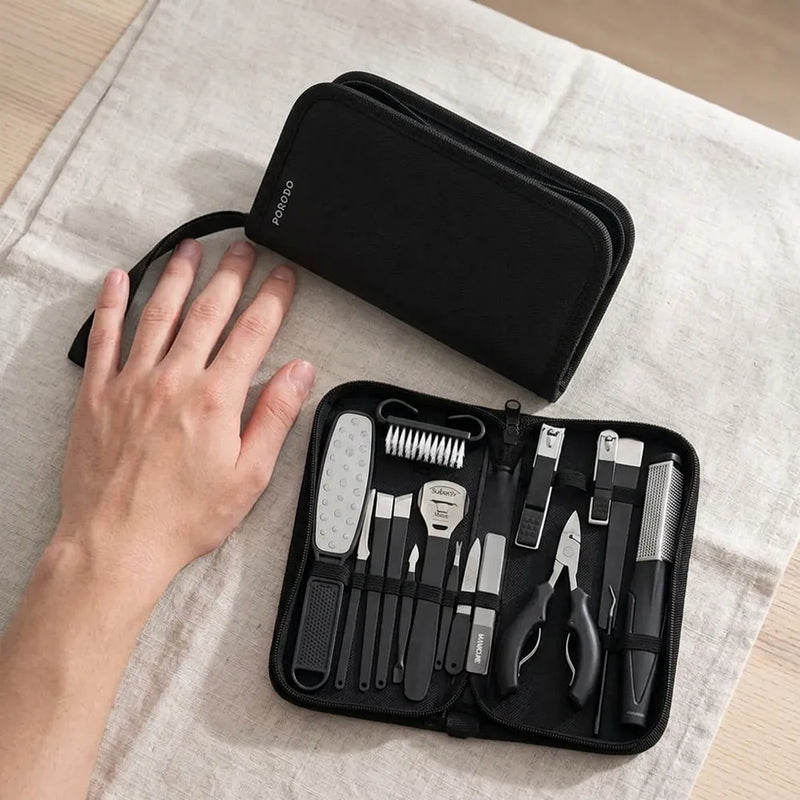 A hand is shown beside an open black zippered nail care set on a light beige fabric surface, with the matching closed case placed above it. The set includes black-handled tools and silver metal tools inside a black organizer case.