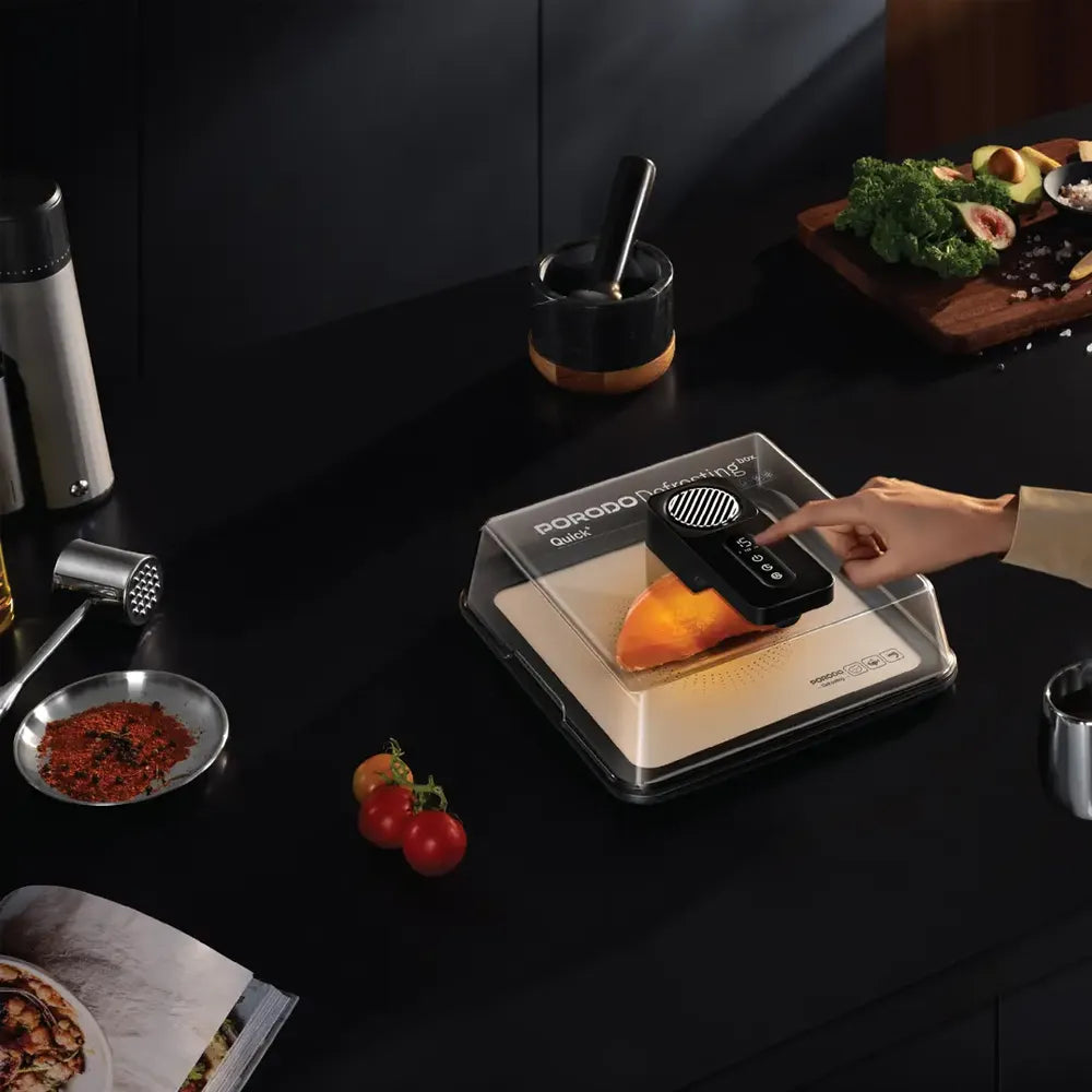 A hand is pressing a black control button on a portable food dehydrator by Porogi, placed on a dark kitchen countertop with food ingredients nearby.