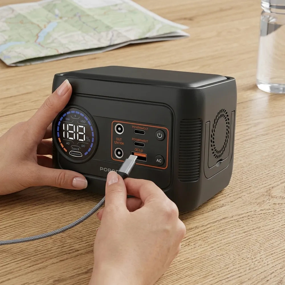 A portable black power bank with a digital display and multiple charging ports, being plugged in by two hands on a wooden table.