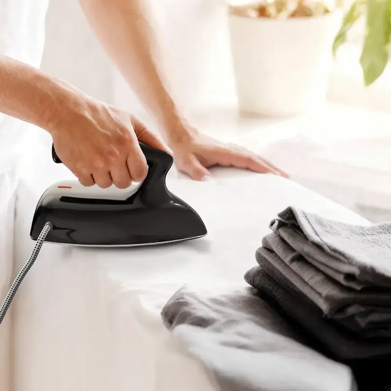 Person using a black Porodo Ironix mini iron on white fabric beside a stack of folded clothes.