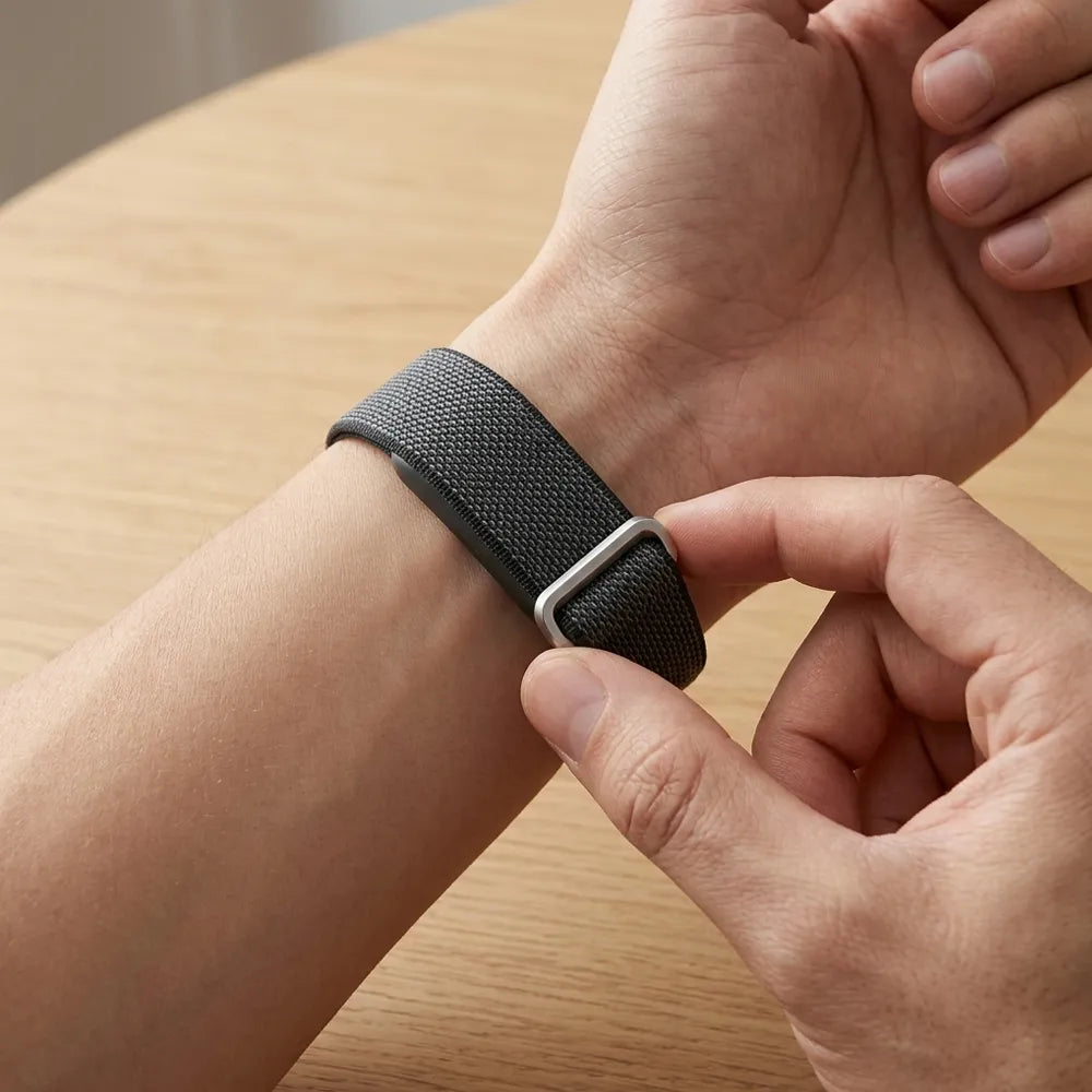 A close-up of a black woven watch band with a silver metal buckle being adjusted on a person's wrist, set against a light wood tabletop background.