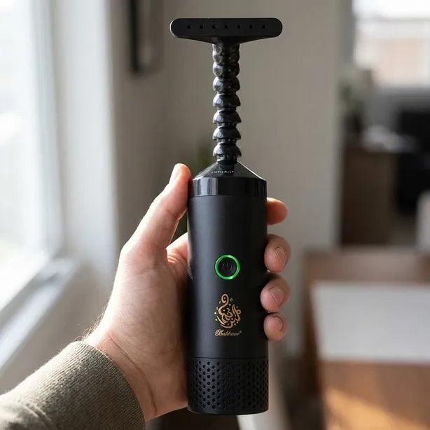 A hand holding a black handheld electric grinder with a textured handle and a green power button, set against a blurred indoor background.