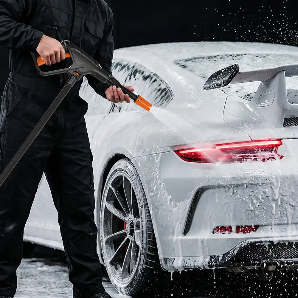 A person in black clothing uses a high-pressure foam cannon to wash a white sports car covered in soap foam, with water spraying and droplets visible against a dark background.